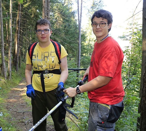 Zwei junge Männer stehen an einem Waldweg und richten sich Werkzeug.