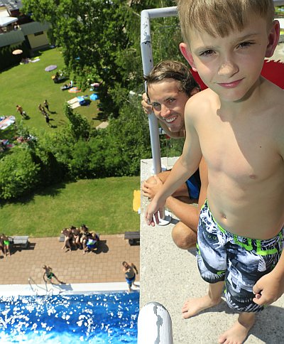 Ein Junge in Badehose steht mit seinem Assistenten am 10 Meter Turm. Unter ihm das Schwimmbad, Liegewiese und Badegäste