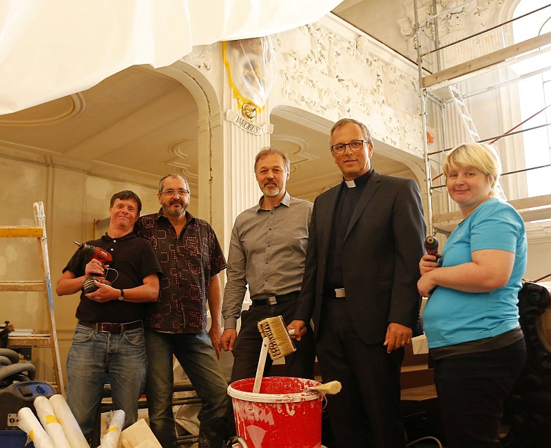 Vier Männer und eine Frau stehen mit Werkzeug in einer Kirche. Gerüste an den Wänden, Schutzfolien auf den Bänken.