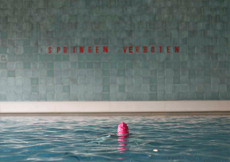 Aus dem Wasser eines Hallen-Schwimmbades schautnur eine Badehaube aus dem Wasser. An der Wand steht "Springen Verboten"