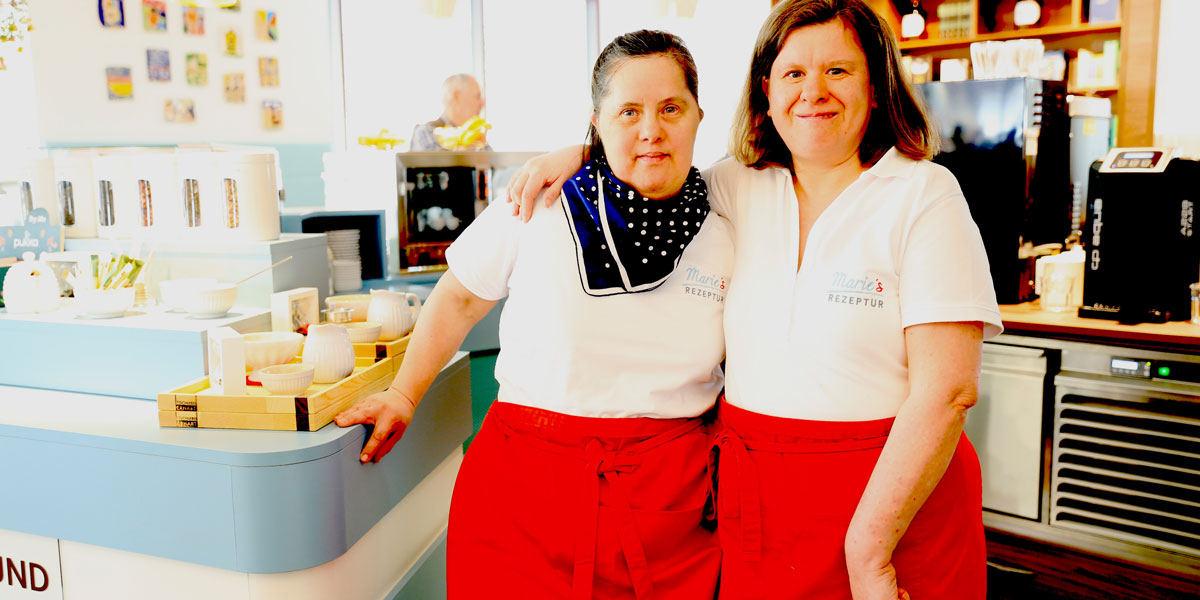 Zwei Servierfachkräfte in einem Cafe lächeln freundlich in die Kamera.
