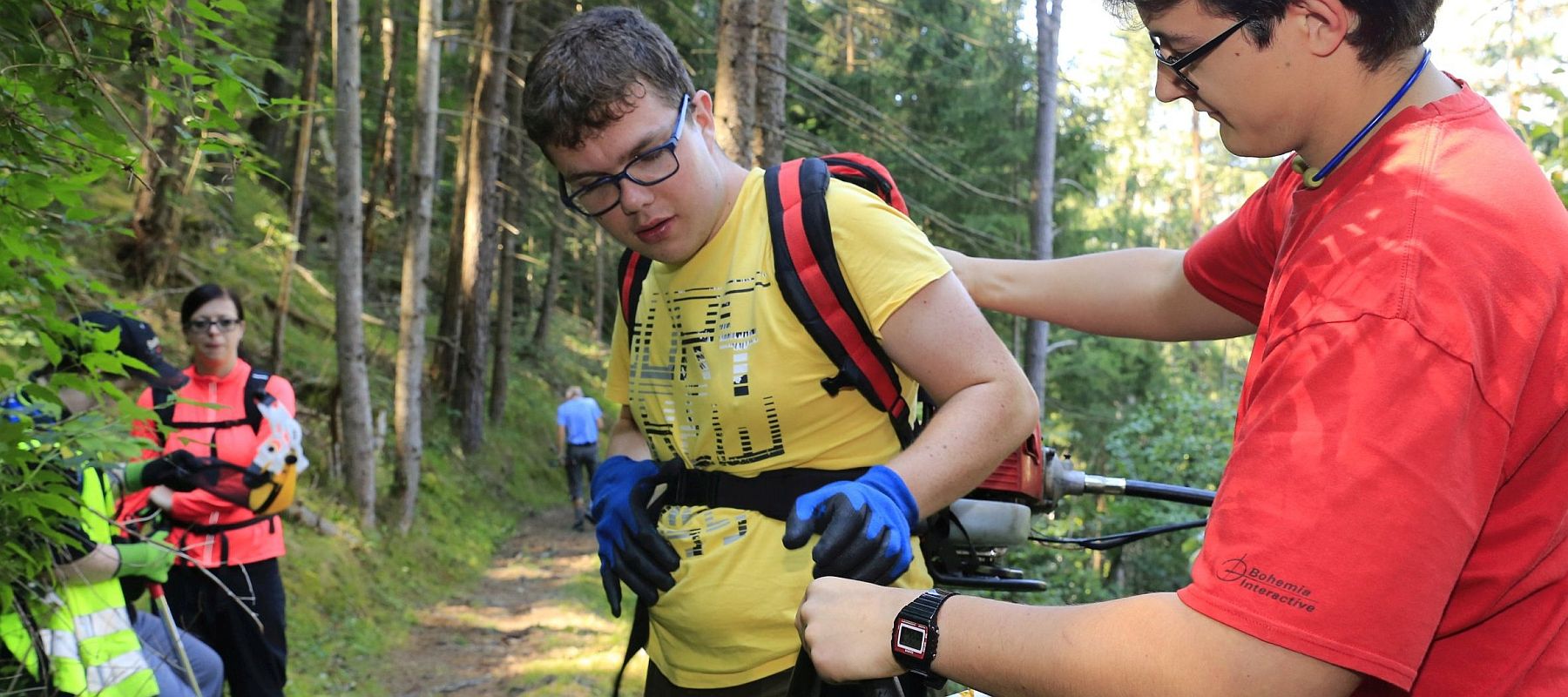 Zwei junge Männer stehen an einem Waldweg und richten sich Werkzeug.