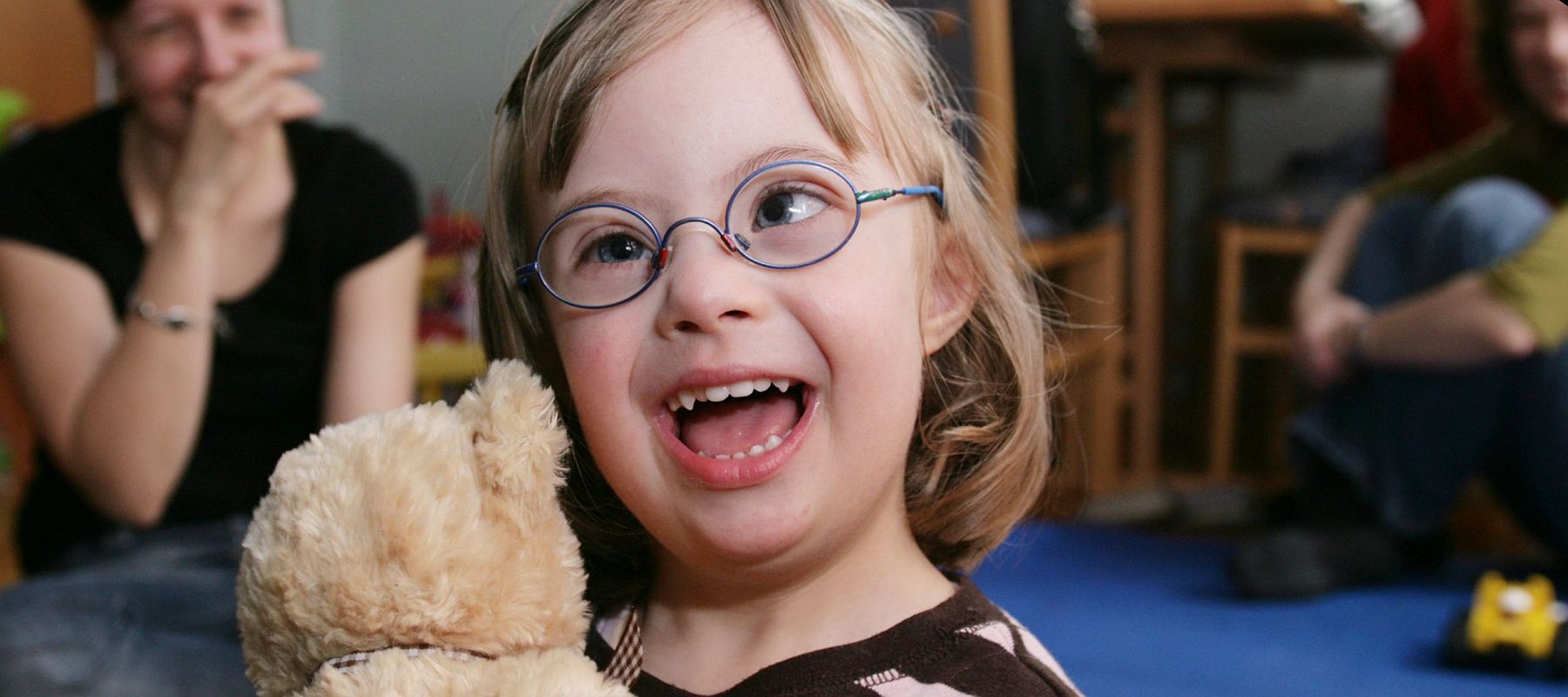 Ein kleines Mädchen sitzt im Kinderzimmer, hält einen Teddybär und lacht. Im Hintergrund sieht man eine Frau.