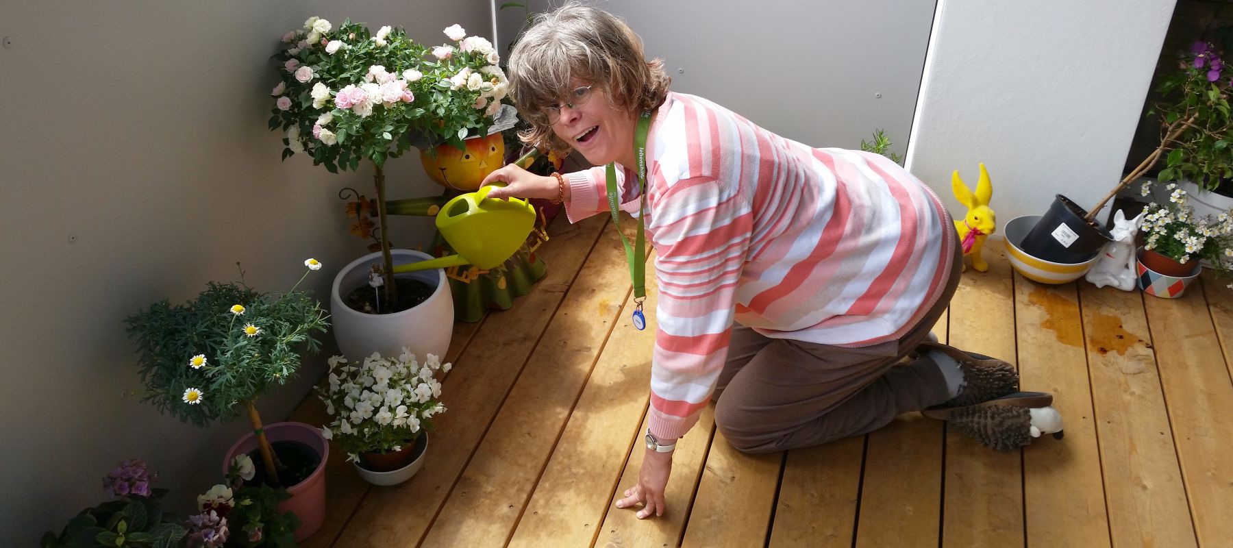 Frau kniet mit einer Gieskanne am Balkon und gießt Blumentöpfe