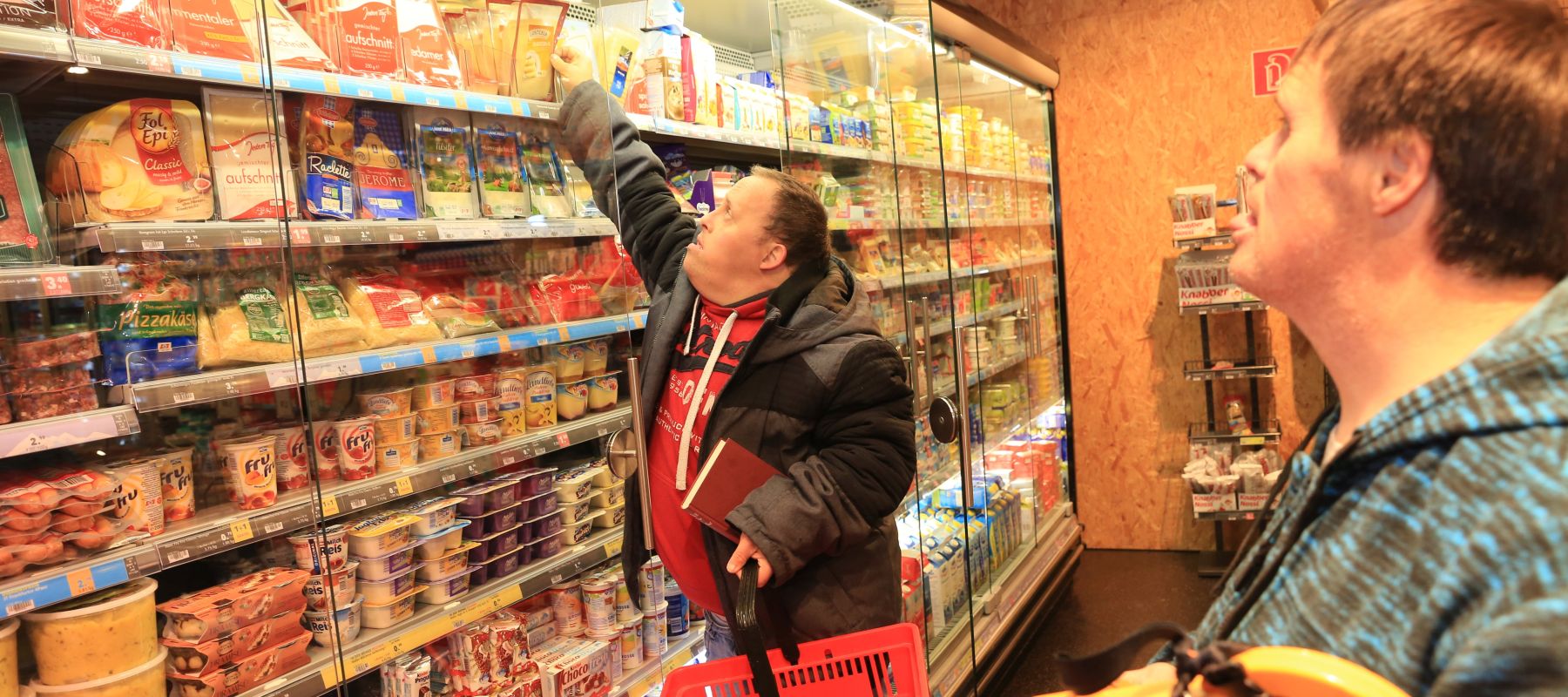 Zwei Männer mit Trisomie 21 stehen vor Supermarkt-Regalen und kaufen ein.