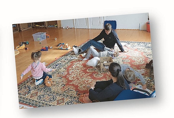 In einen großen Zimmer spielen drei kleine Kinder am Teppich. Zwei Erwachsene beaufsichtigen sie.