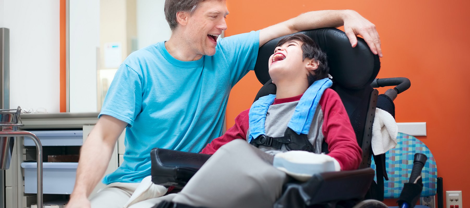 Ein Vater spricht und lacht mit seinem Kind. Der Sohn sitzt im Rollstuhl und lacht.