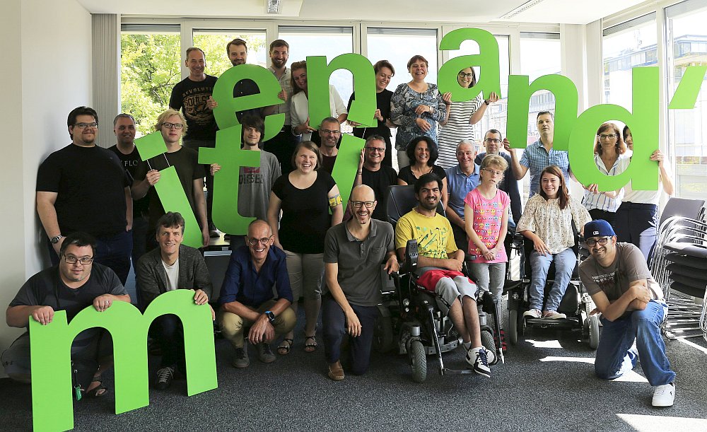 30 gutgelaunte Menschen mit und ohne Behinderungen halten Buchstaben, die das Wort "miteinand'" ergeben.